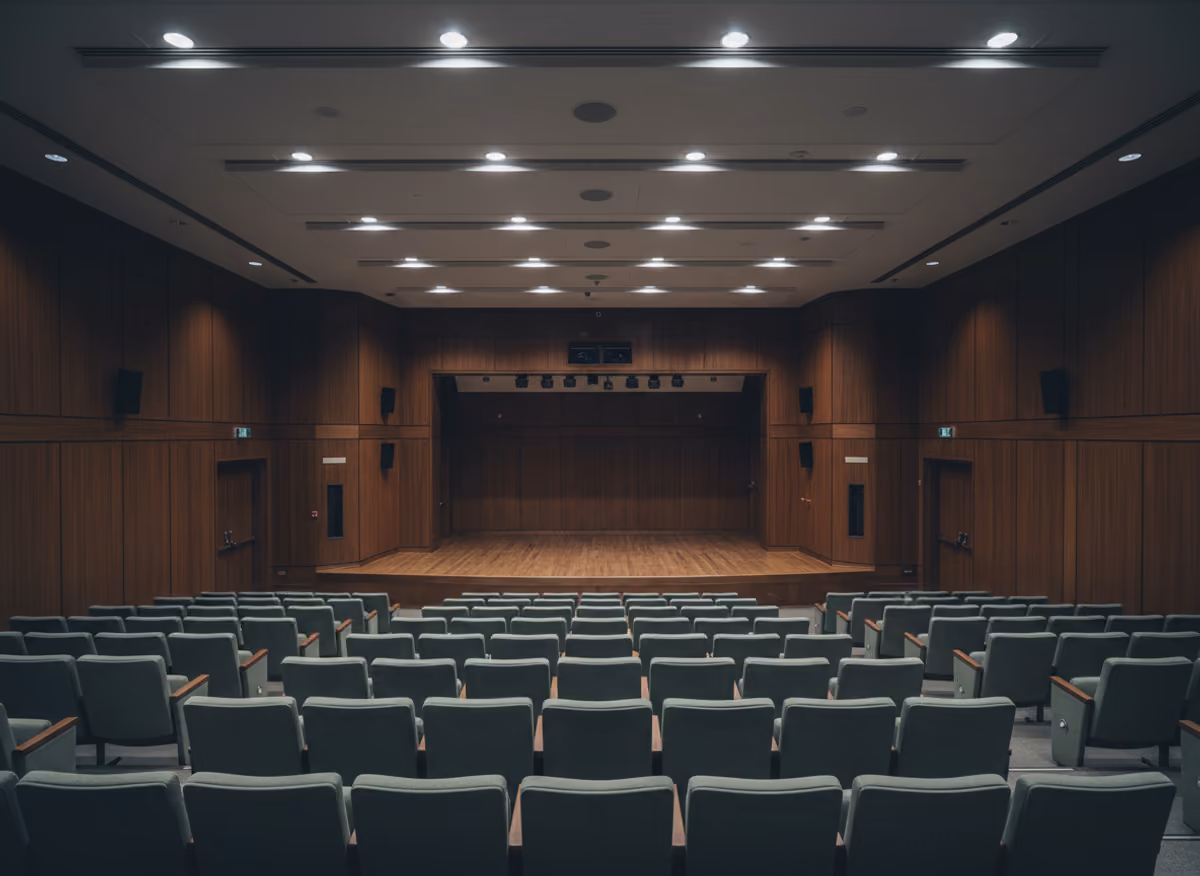 Empty raked auditorium seating facing a low stage, warm wood panelling, house lights down.