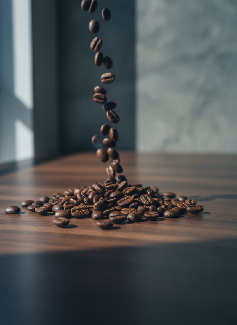 Whole roasted coffee beans spilled across a dark walnut counter in cold window light.