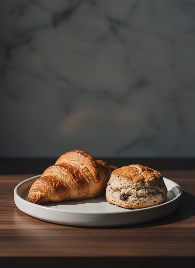 A butter croissant and a scone on a pale ceramic plate on a dark walnut counter.